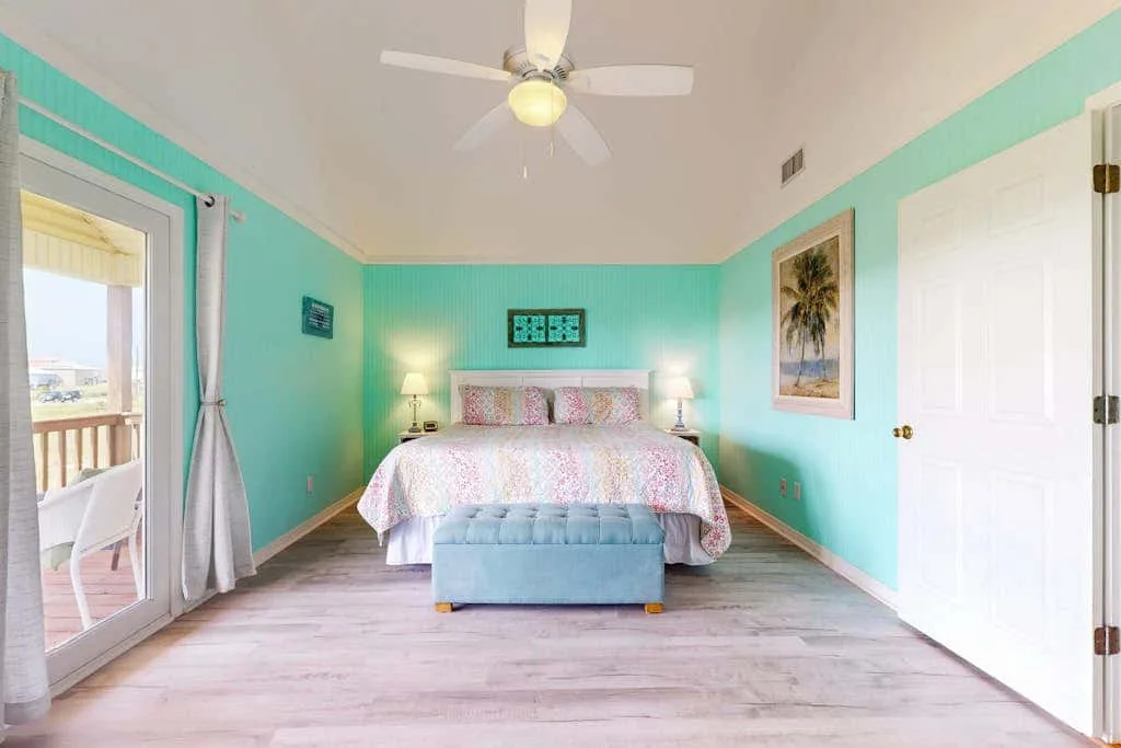 A bright bedroom with a bed, turquoise walls, and glass doors opening to a balcony.