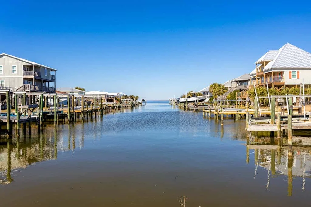 A calm waterway runs between waterfront homes and docks under a clear blue sky.