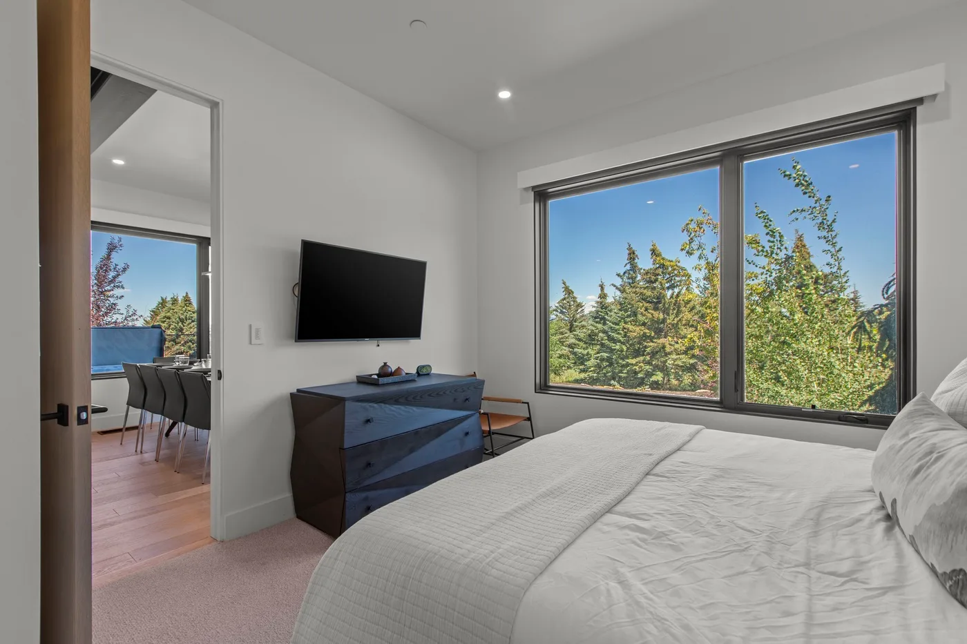 Un dormitorio luminoso con una cama, un televisor y una gran ventana con vista a los árboles.
