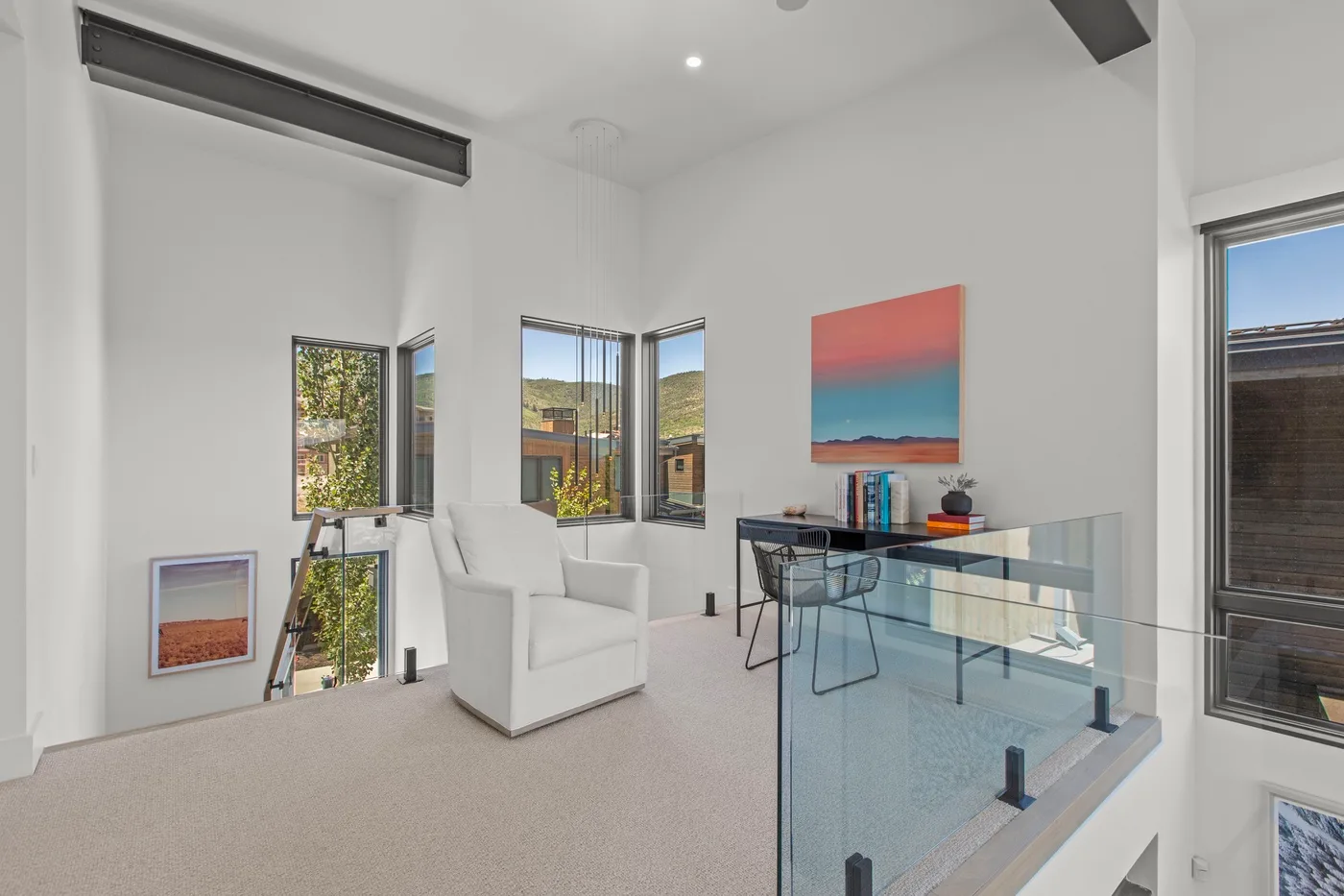 Una luminosa sala de estar tipo loft con un sillón blanco, barandilla de vidrio y grandes ventanas.