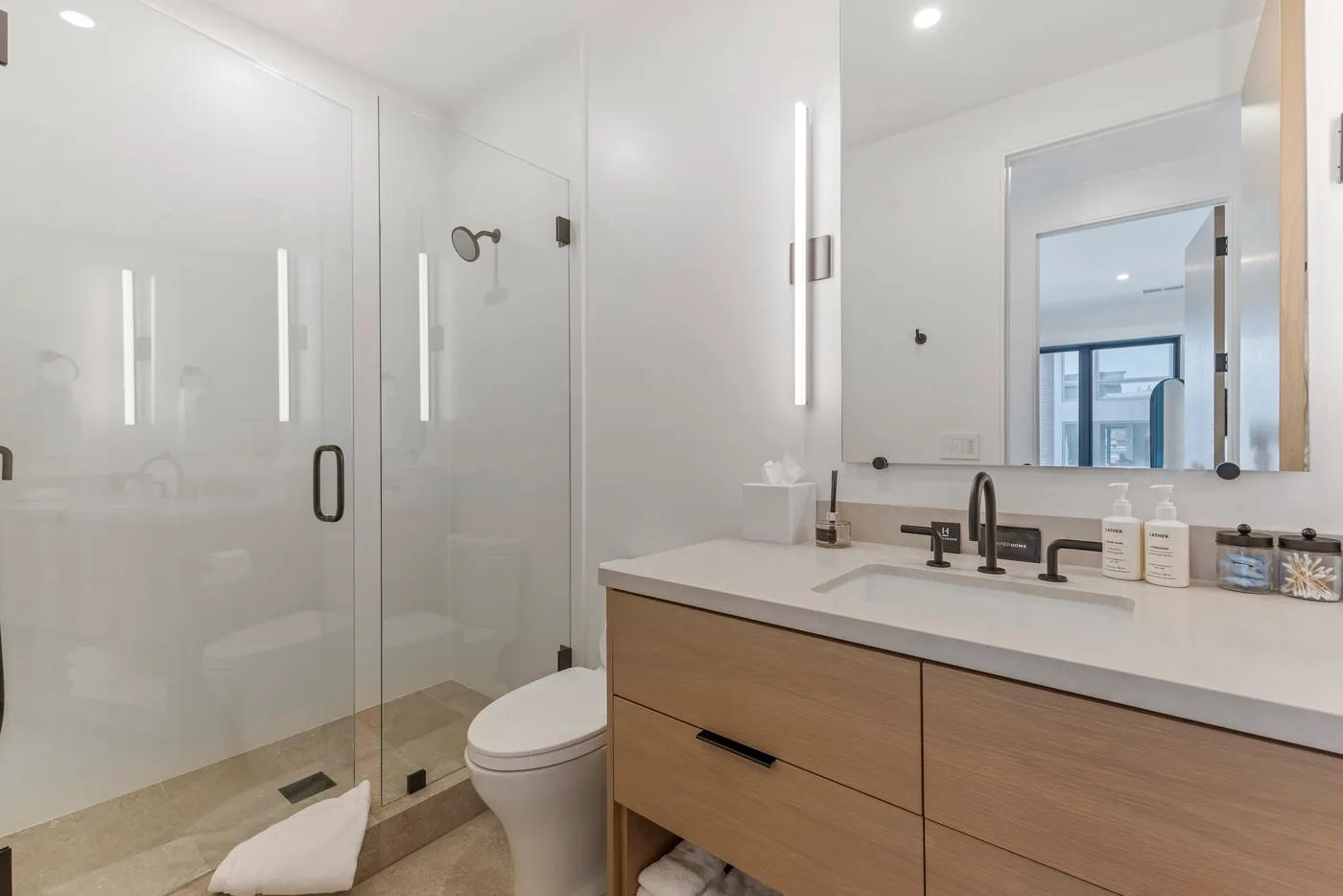 Baño luminoso con ducha de vidrio, inodoro y tocador de madera.