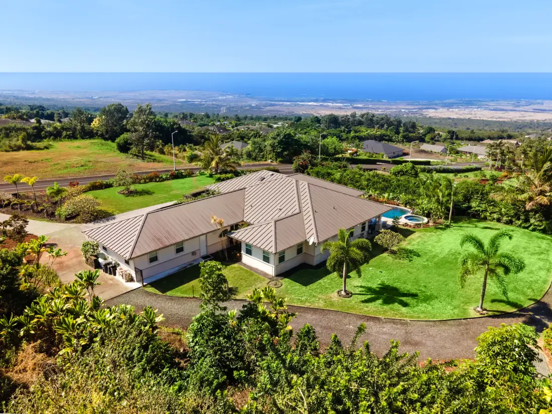 Vista aérea de una casa con piscina entre jardines verdes y el océano a lo lejos.