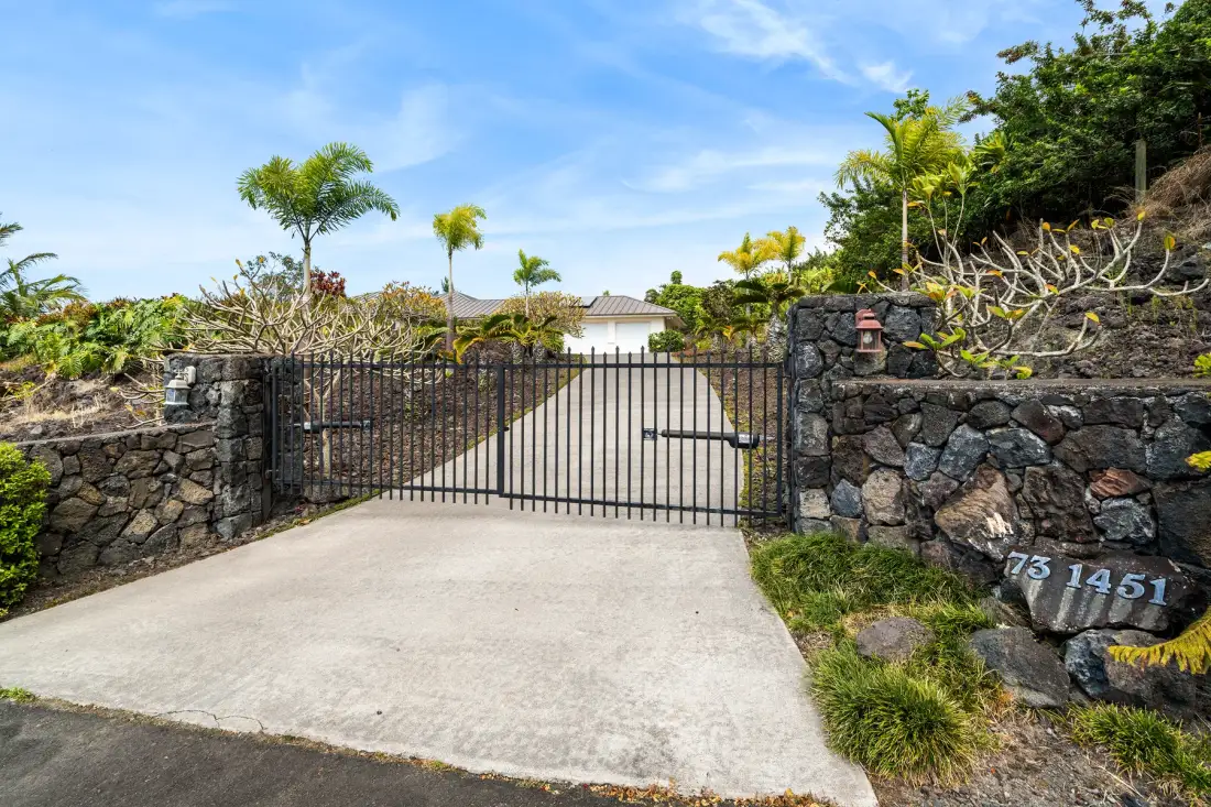Una entrada cerrada conduce hacia un edificio, flanqueada por muros de piedra y plantas tropicales.