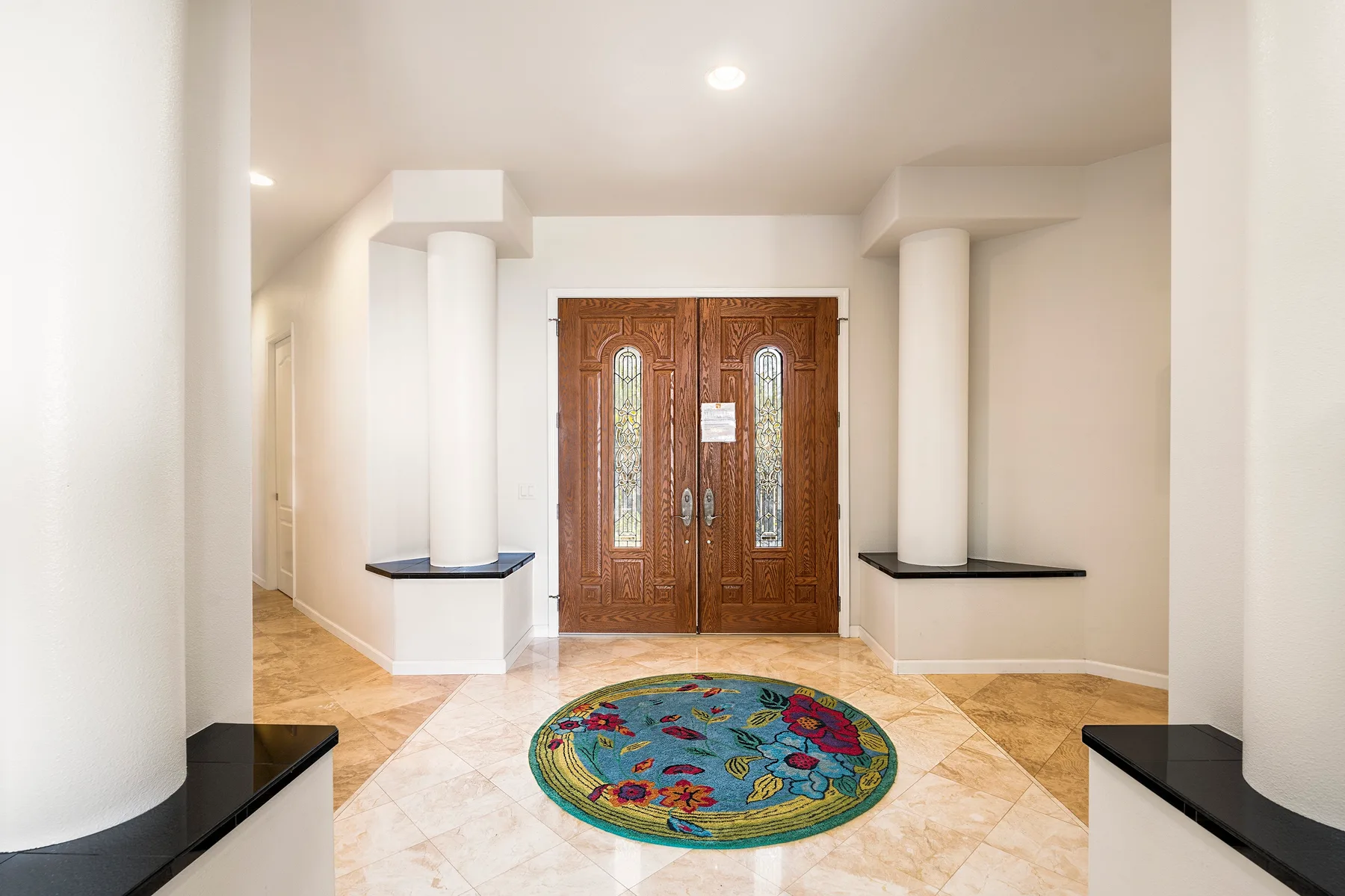 Un recibidor luminoso presenta una puerta doble de madera ornamentada y una alfombra redonda colorida.
