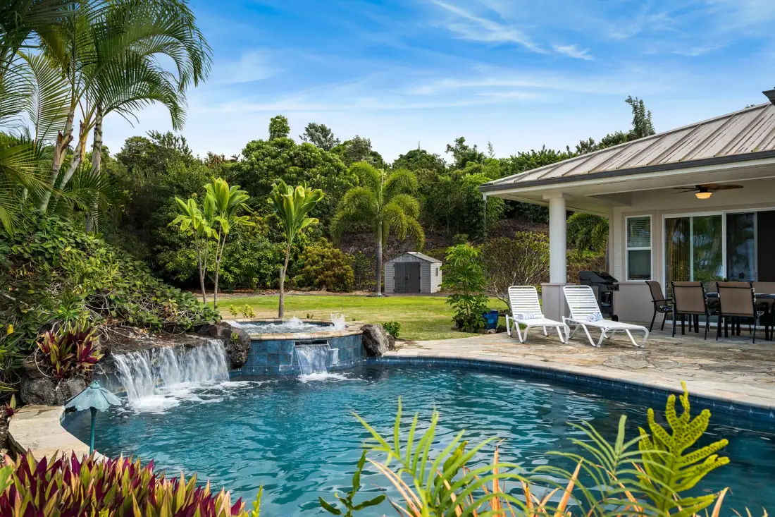 Una piscina al aire libre con cascada junto a un patio cubierto, rodeada de plantas tropicales.