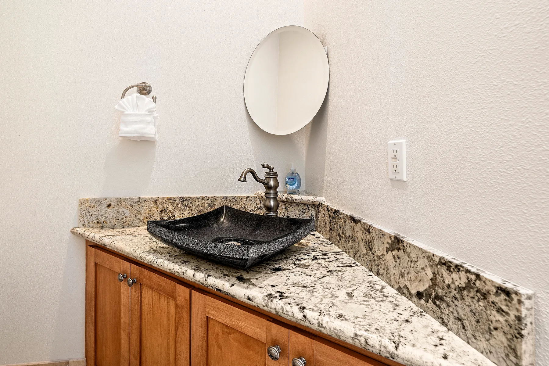 Lavabo de baño con un lavabo oscuro sobre encimera y un espejo redondo.