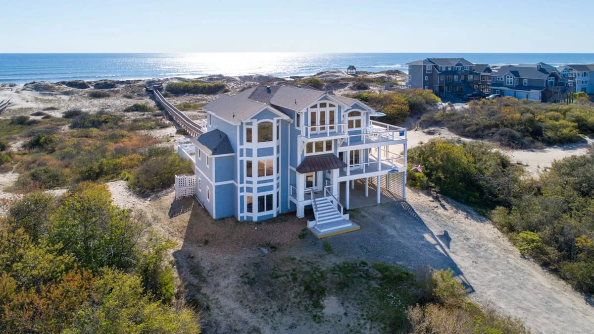A large coastal house stands among dunes beside the ocean.