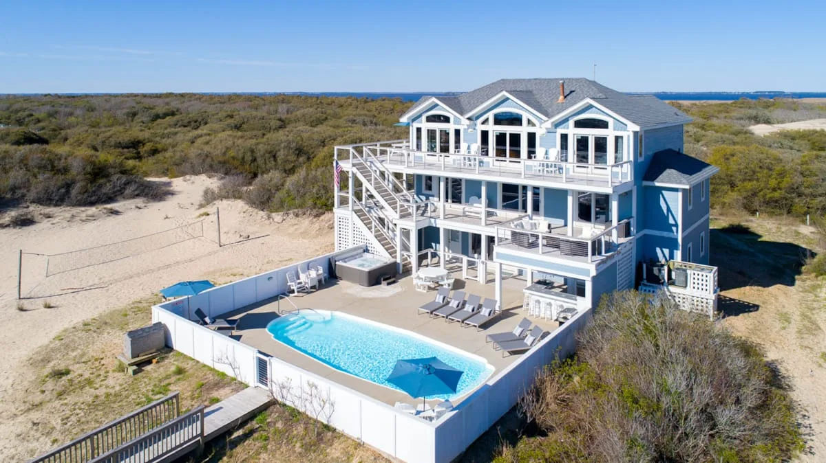 A large coastal house overlooks a fenced swimming pool and sandy dunes.