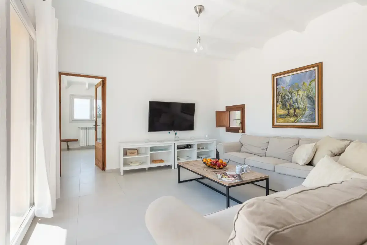 Salón luminoso con asientos claros, un televisor y una mesa de centro.