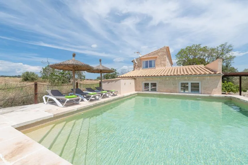 Una piscina junto a una casa de techo de tejas, con tumbonas y sombrillas.