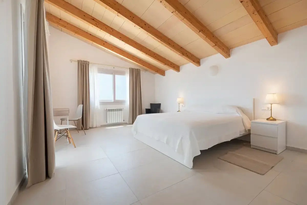 Un dormitorio luminoso con una cama, mesillas de noche y vigas vistas en el techo.
