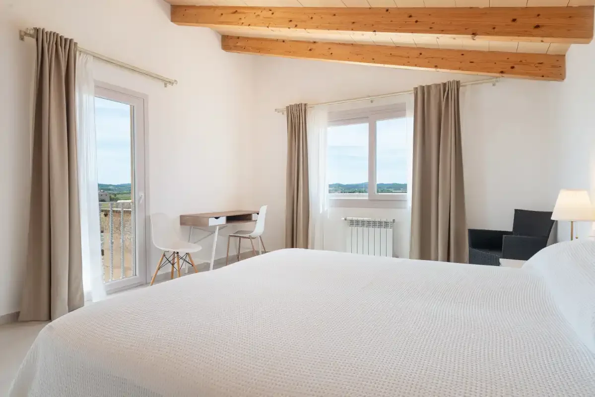 Un dormitorio luminoso con una cama, una mesa pequeña y ventanas con vistas al paisaje.