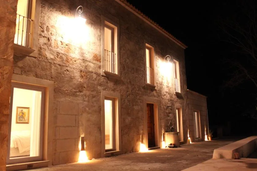 Exterior de un edificio de piedra por la noche, iluminado por luces de pared y ventanas encendidas.