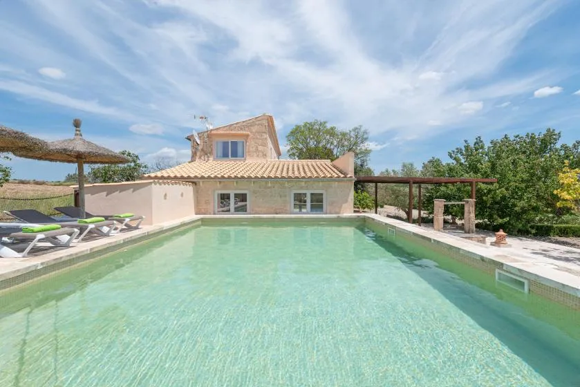 Una piscina exterior está junto a una casa con techo de tejas y una zona de sombra.