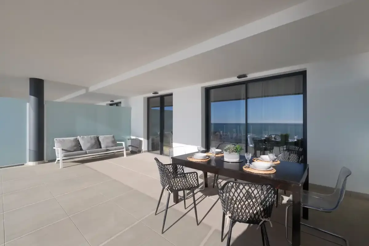 Una terraza cubierta tiene una mesa de comedor y asientos junto a ventanas con vistas al mar.