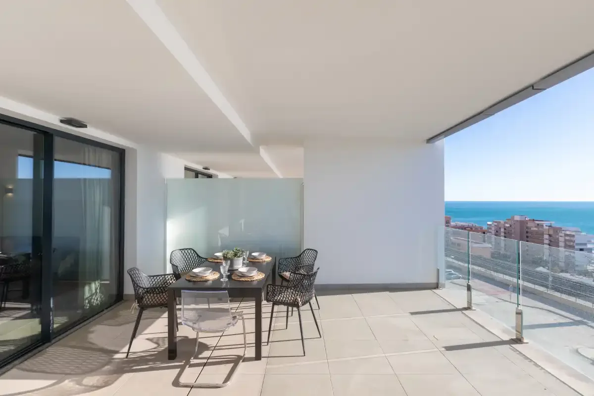 Una terraza con suelo de baldosas tiene una mesa de comedor y asientos junto a una barandilla de cristal con vistas al mar.