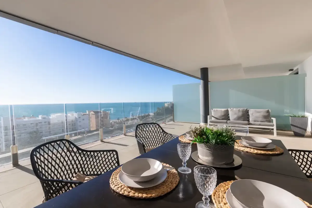 Una terraza con vistas al mar, muebles de comedor exterior y una barandilla de vidrio.