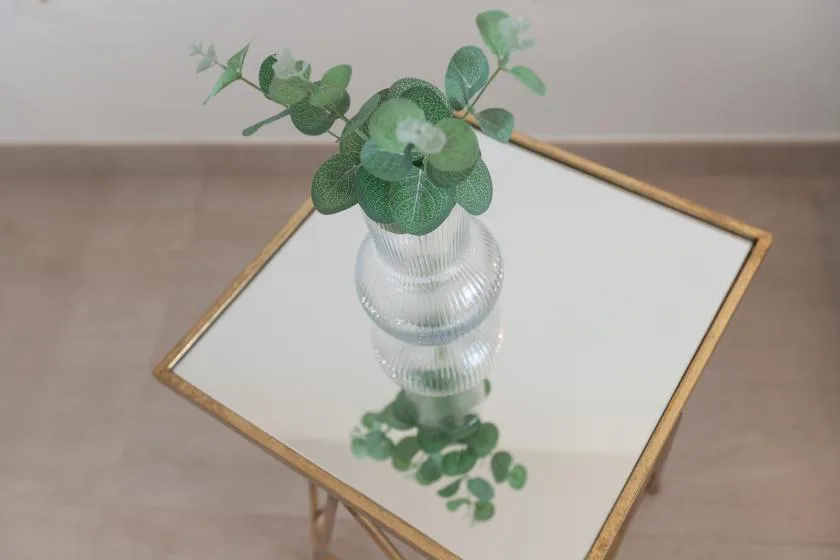 Un jarrón de vidrio con follaje verde está sobre una mesa con espejo.