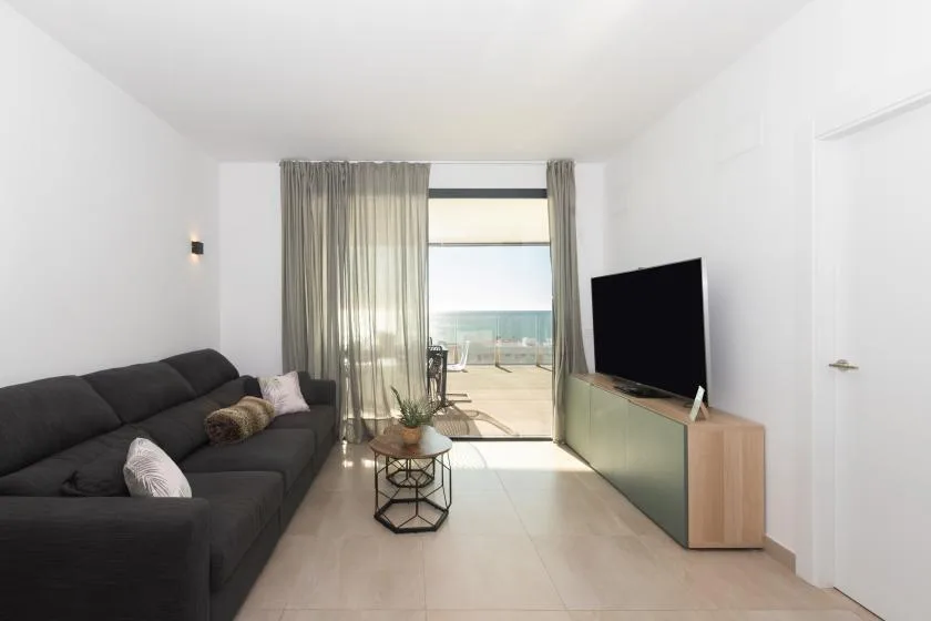 Una sala luminosa con asientos oscuros, un televisor y puertas de vidrio que dan a un balcón.