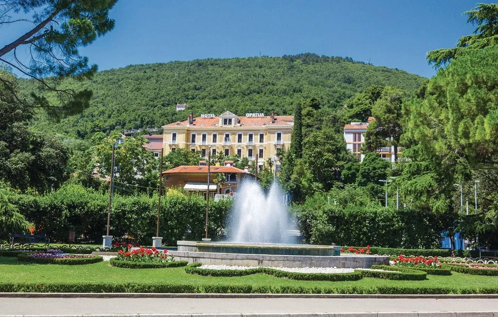 A fountain sprays water in a landscaped garden before a large building and wooded hillside.