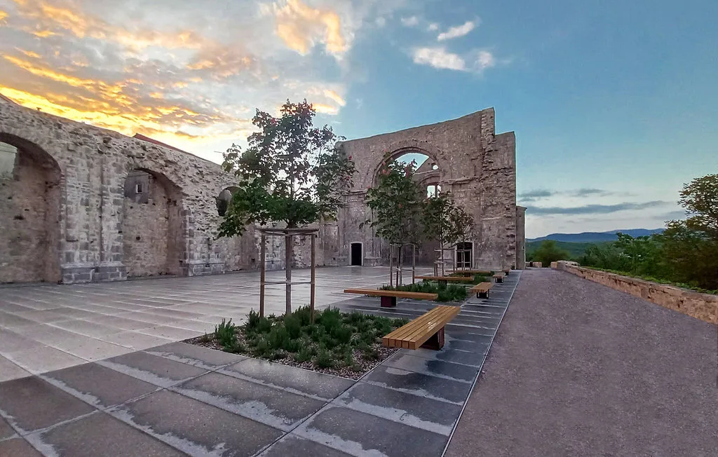 An open stone courtyard with ruined walls, benches, and trees beneath a colorful evening sky.