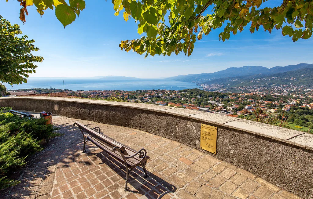 A stone-paved lookout terrace has a bench and a view of the coastal town and mountains.