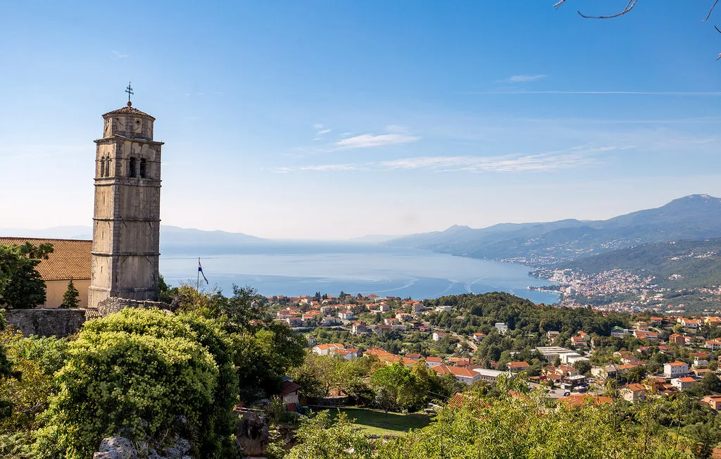 A scenic hillside town overlooks a broad lake beneath a clear blue sky.