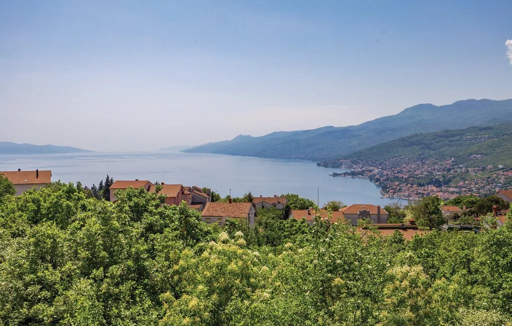 A lakeside town and distant mountains are seen beyond green trees.