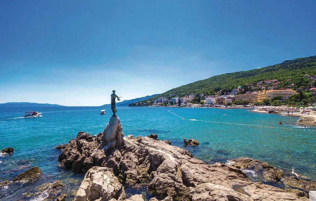 A rocky shoreline extends into turquoise water, with a statue and coastal buildings beyond.