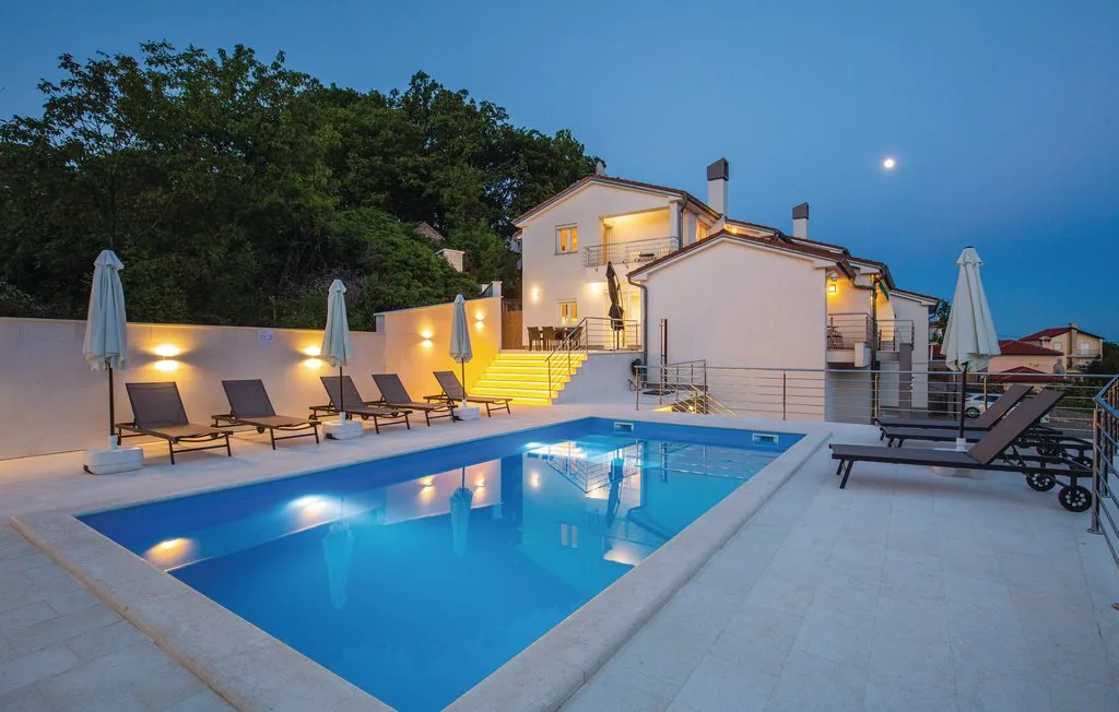 Una piscina al aire libre junto a una casa iluminada y asientos al anochecer.