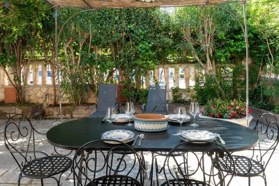 Una mesa de comedor al aire libre está puesta bajo una pérgola frondosa.
