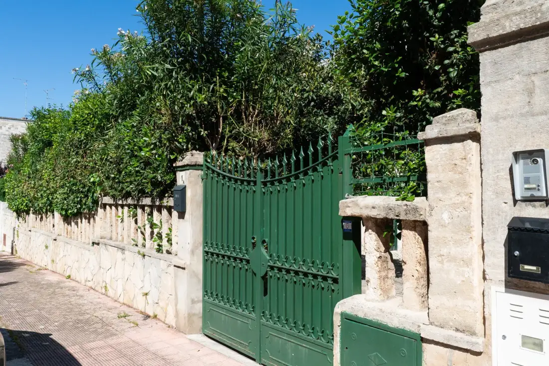 Una puerta metálica verde y un muro de piedra clara bordean un camino exterior arbolado.