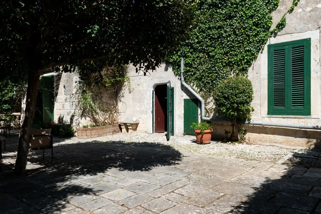Un patio de piedra a la sombra de un árbol frondoso, con contraventanas verdes en el edificio.