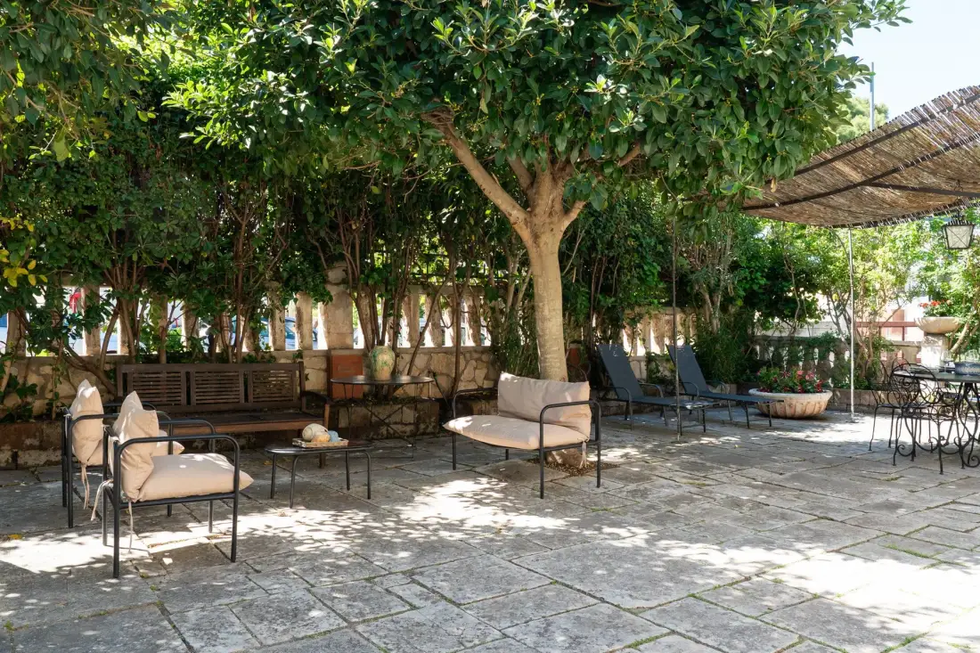 Una zona de estar al aire libre a la sombra de árboles frondosos, con mesas y sillas en un patio pavimentado.