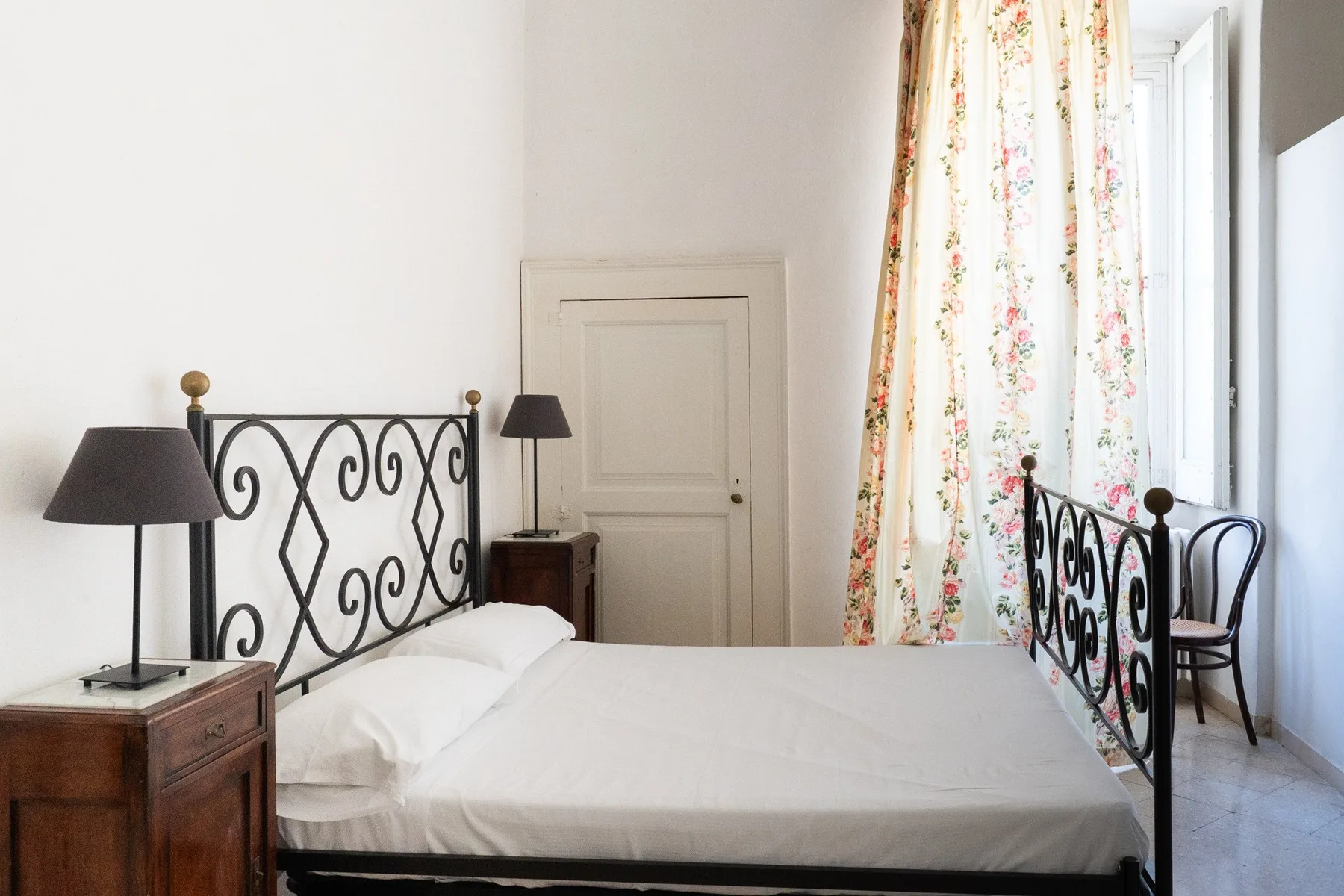 Un dormitorio con una cama blanca, estructura negra de metal y cortinas florales.