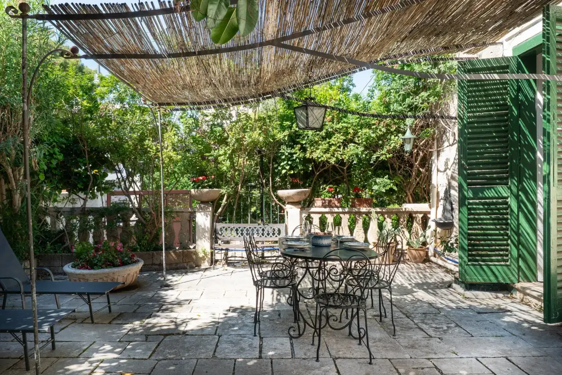 Un patio exterior sombreado tiene una mesa de metal y asientos entre plantas frondosas.