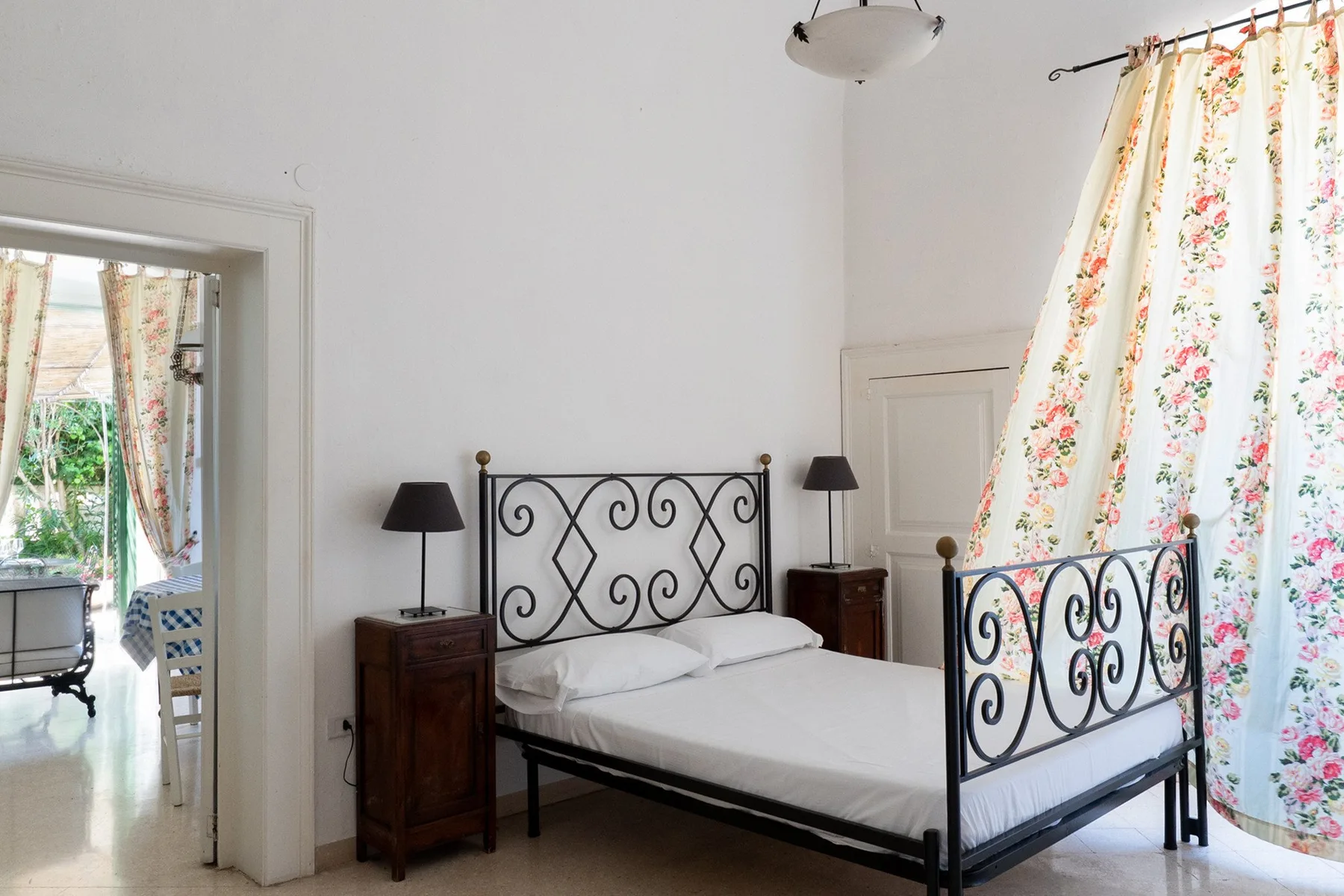 Un dormitorio con una cama de metal negro, ropa de cama blanca y cortinas florales.