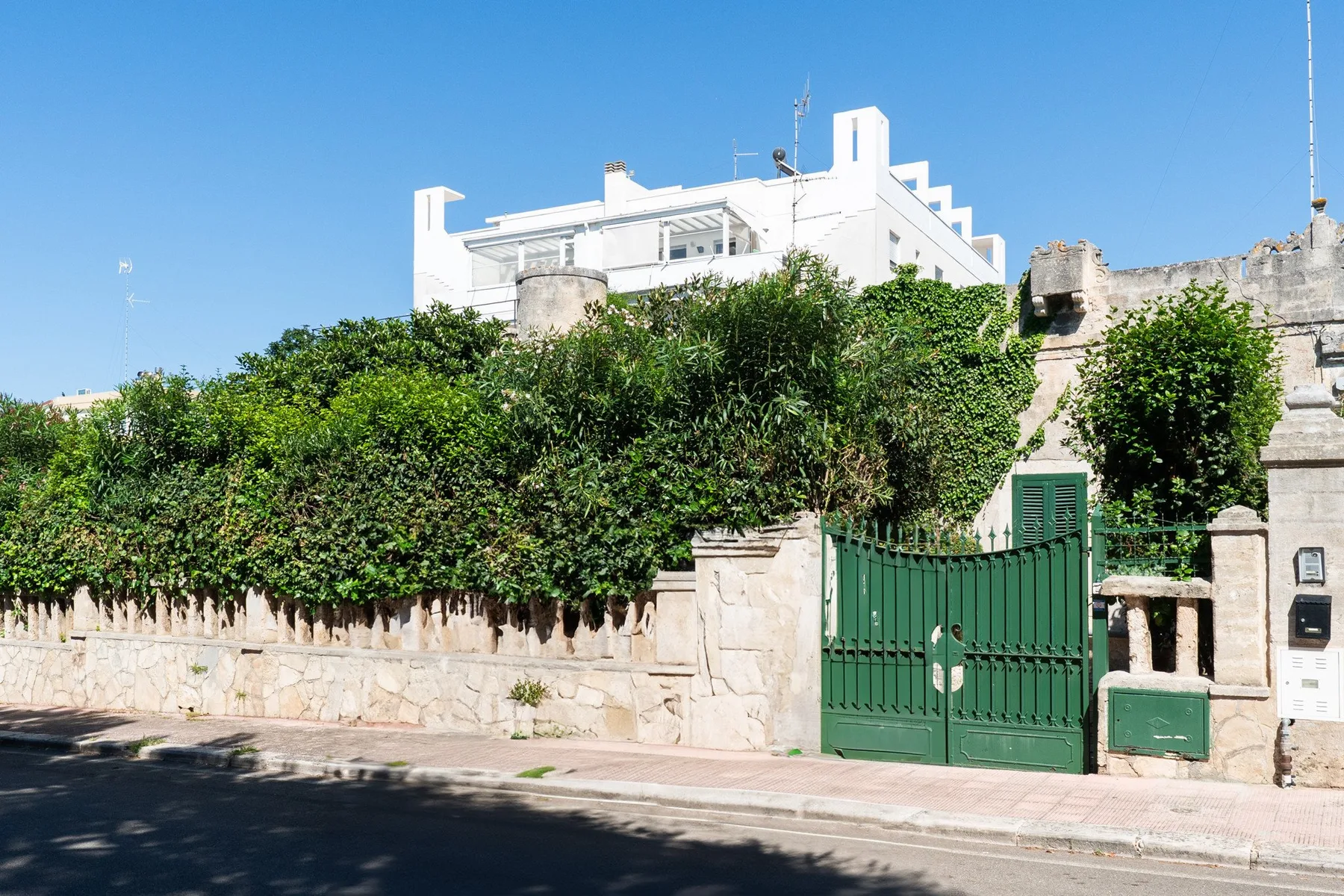 Una verja metálica verde y un seto frondoso bordean un muro de piedra junto a una calle soleada.