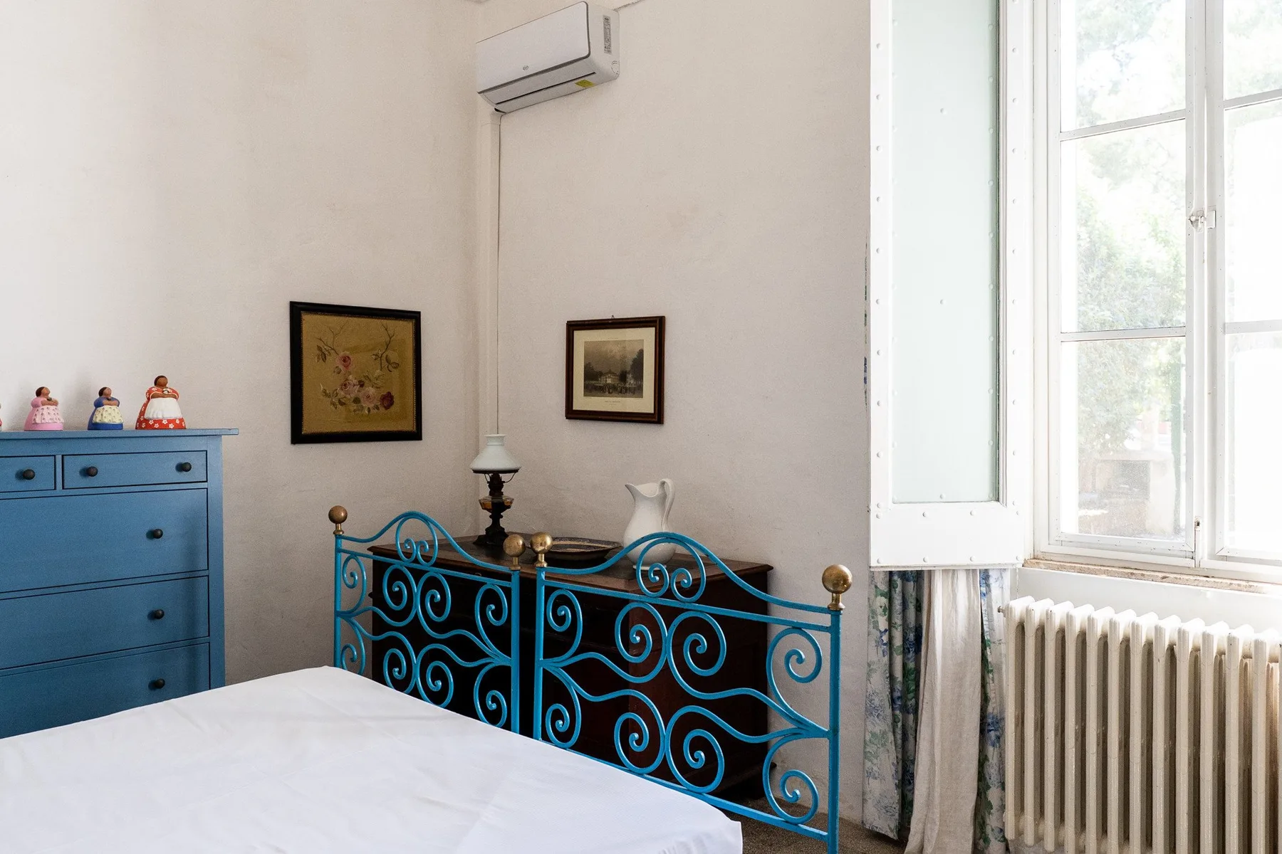 Un dormitorio con cama, cabecero metálico azul, cómoda y ventanas altas.