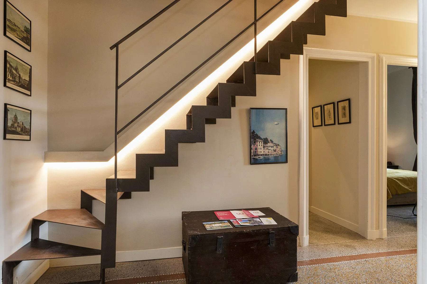 Una escalera con peldaños oscuros y barandilla metálica recorre una pared con cuadros.