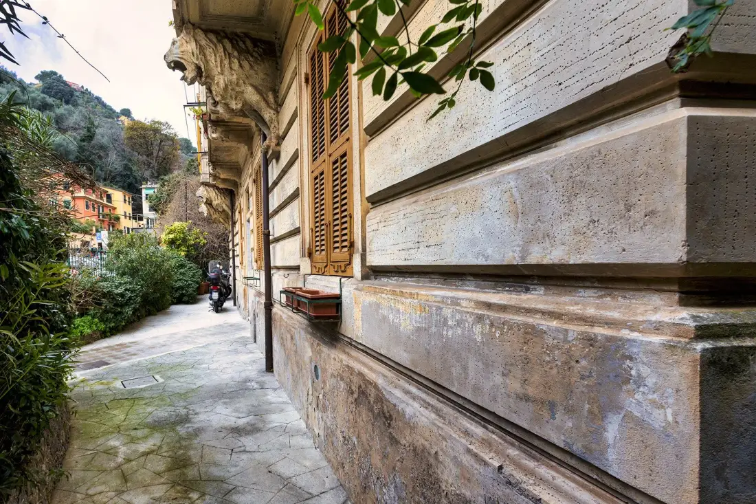 Un camino pavimentado bordea un edificio de piedra con ventanas con contraventanas y plantas frondosas.