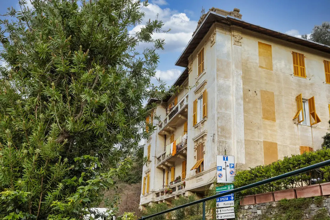 Un edificio antiguo de varias plantas con ventanas con postigos está junto a árboles frondosos.