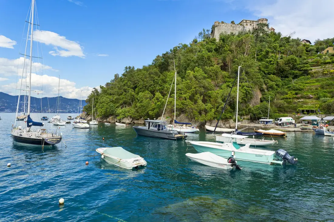 Barcos están amarrados en un puerto costero bajo una colina verde coronada por ruinas de piedra.