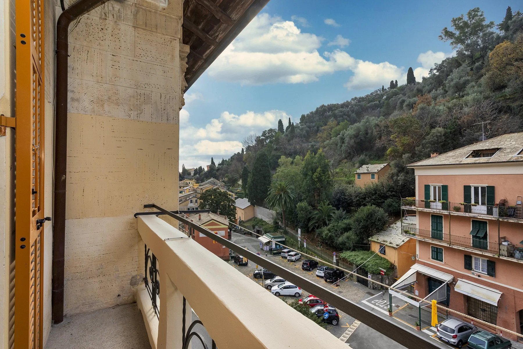 Un balcón da a una calle, edificios cercanos y una ladera verde.