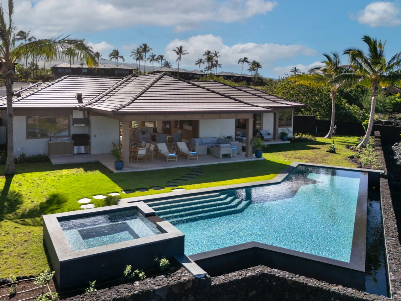 Una casa tropical da a una piscina, con asientos al aire libre bajo el techo.