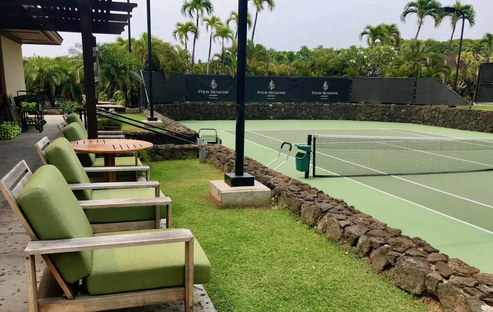 Una cancha de tenis junto a una zona de asientos con césped y palmeras al fondo.