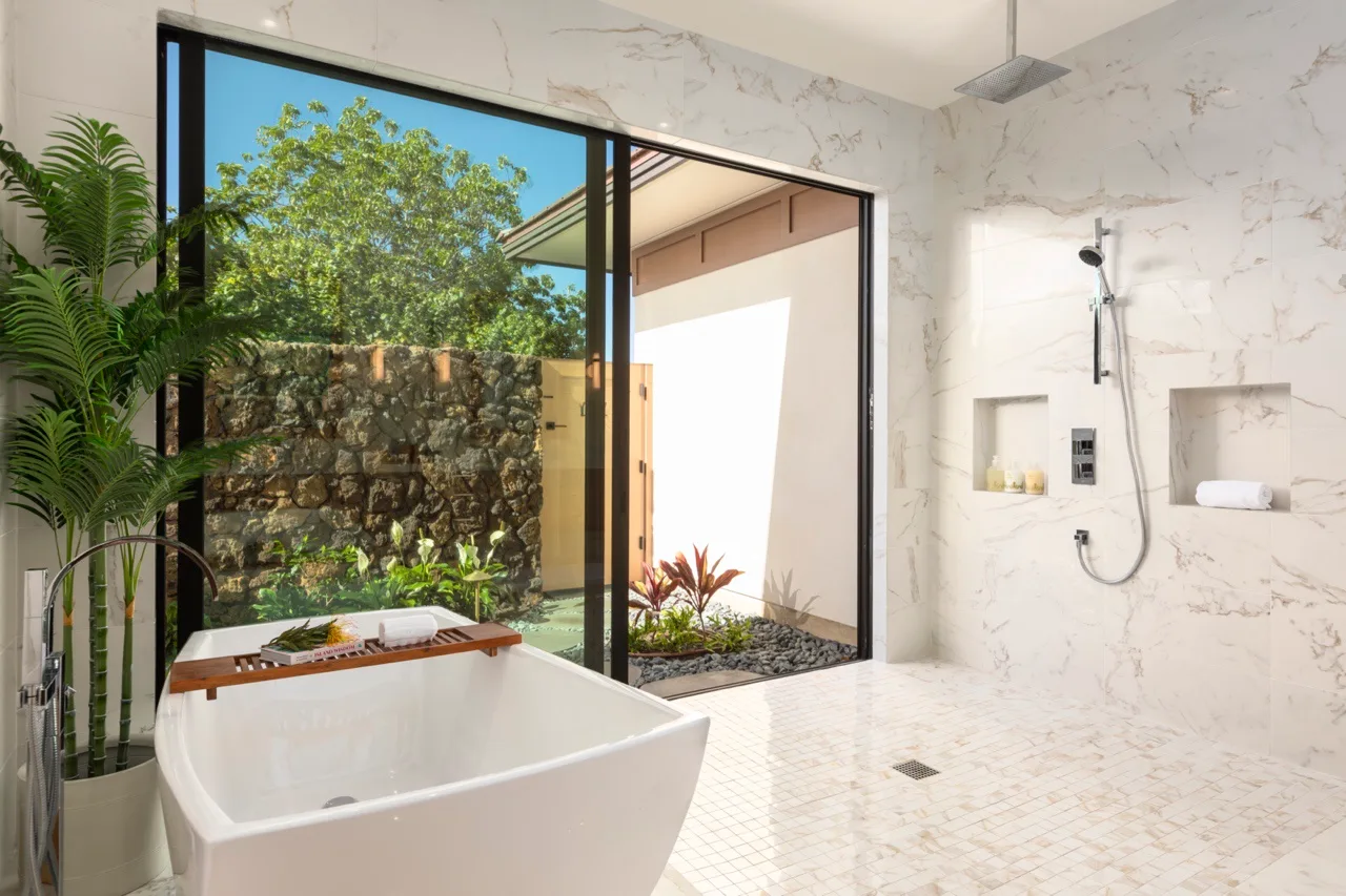Un baño luminoso con bañera, ducha y grandes puertas de vidrio abiertas hacia la vegetación.