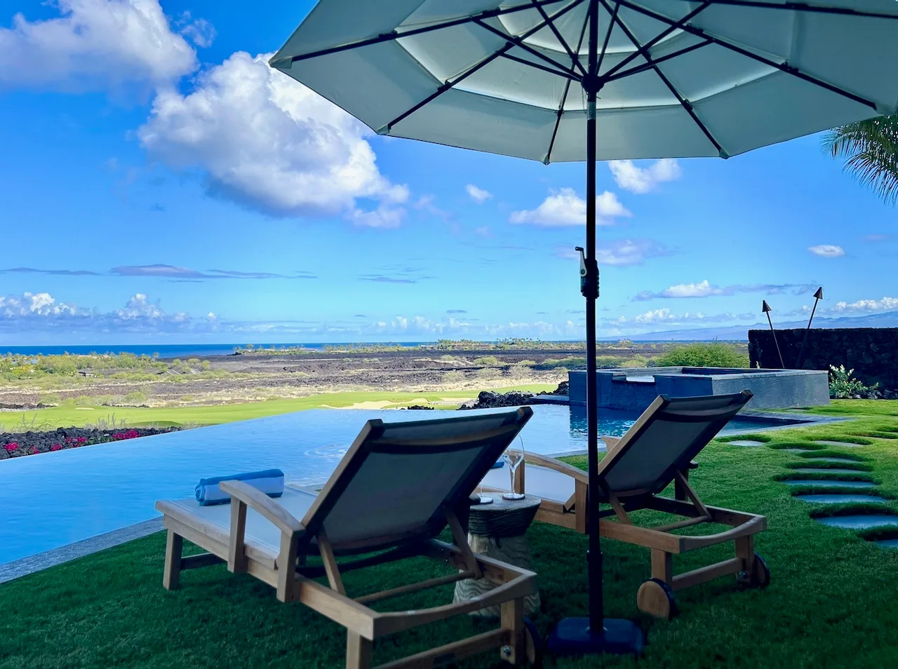Asientos al aire libre junto a una piscina ofrecen vistas de un paisaje costero bajo una sombrilla grande.