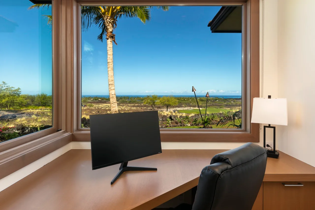 Un escritorio con monitor y asiento mira por grandes ventanas hacia un paisaje costero.