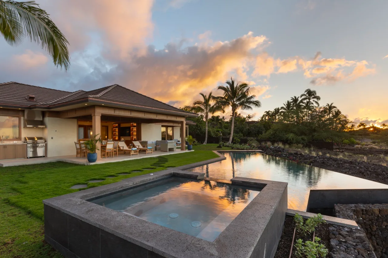 Una casa moderna junto a una piscina al atardecer, con palmeras al fondo.